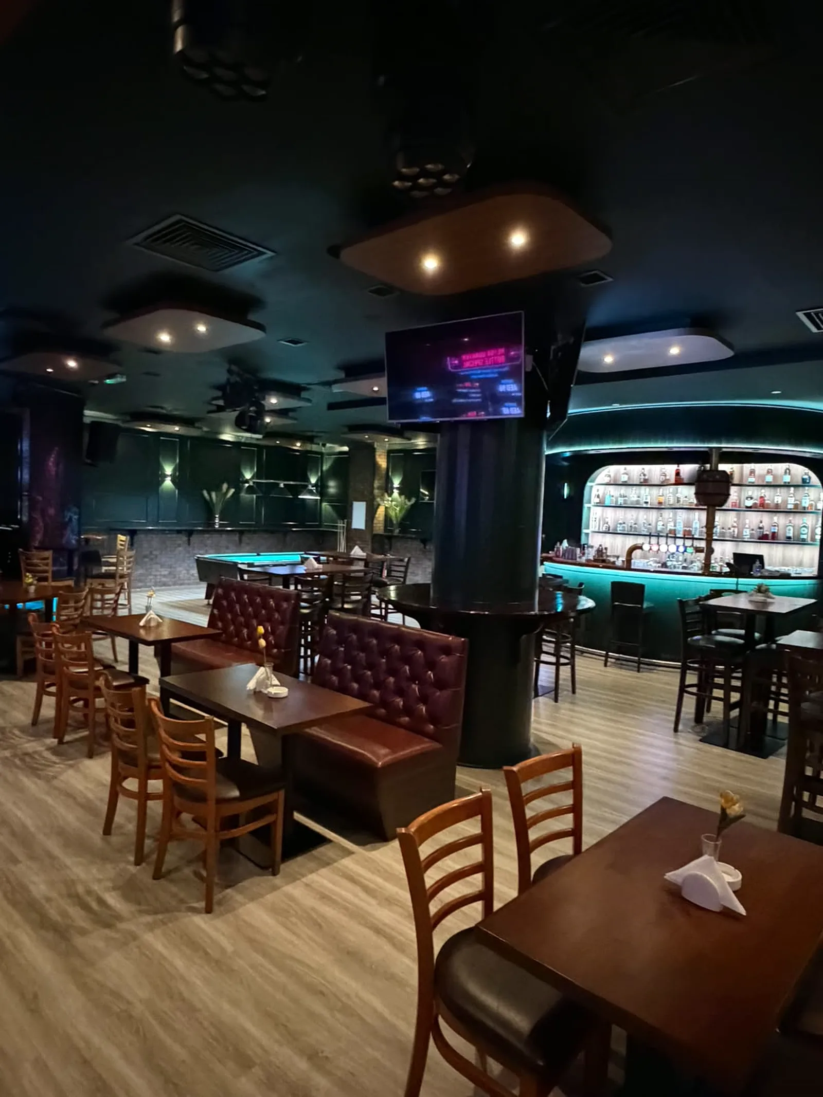 Booth seating around a clad column with mounted screen, leading to the illuminated bar and pool table zone